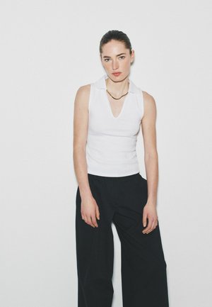 Frau mit zurückgebundenem Haar, die ein ärmelloses weißes Oberteil mit Kragen und lockere schwarze Hosen trägt, steht vor einem schlichten weißen Hintergrund.