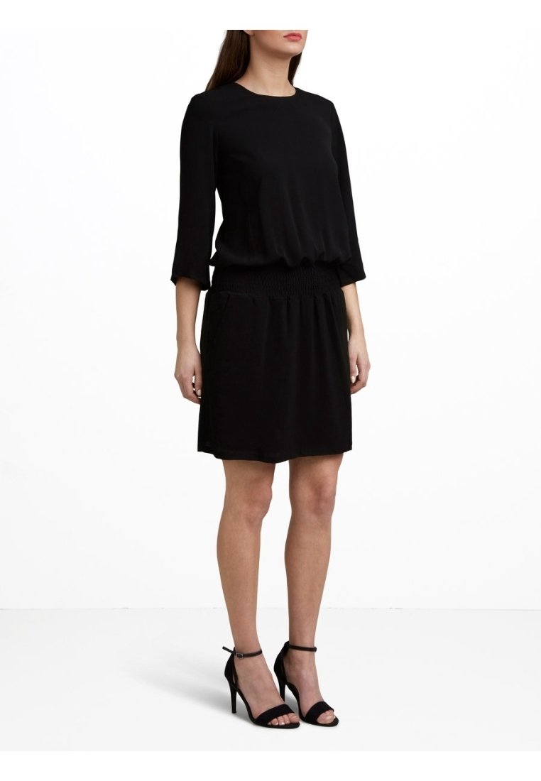 Femme portant une robe noire longueur genou avec manches 3/4 et des sandales noires à talons hauts et brides à la cheville, debout sur un fond blanc.