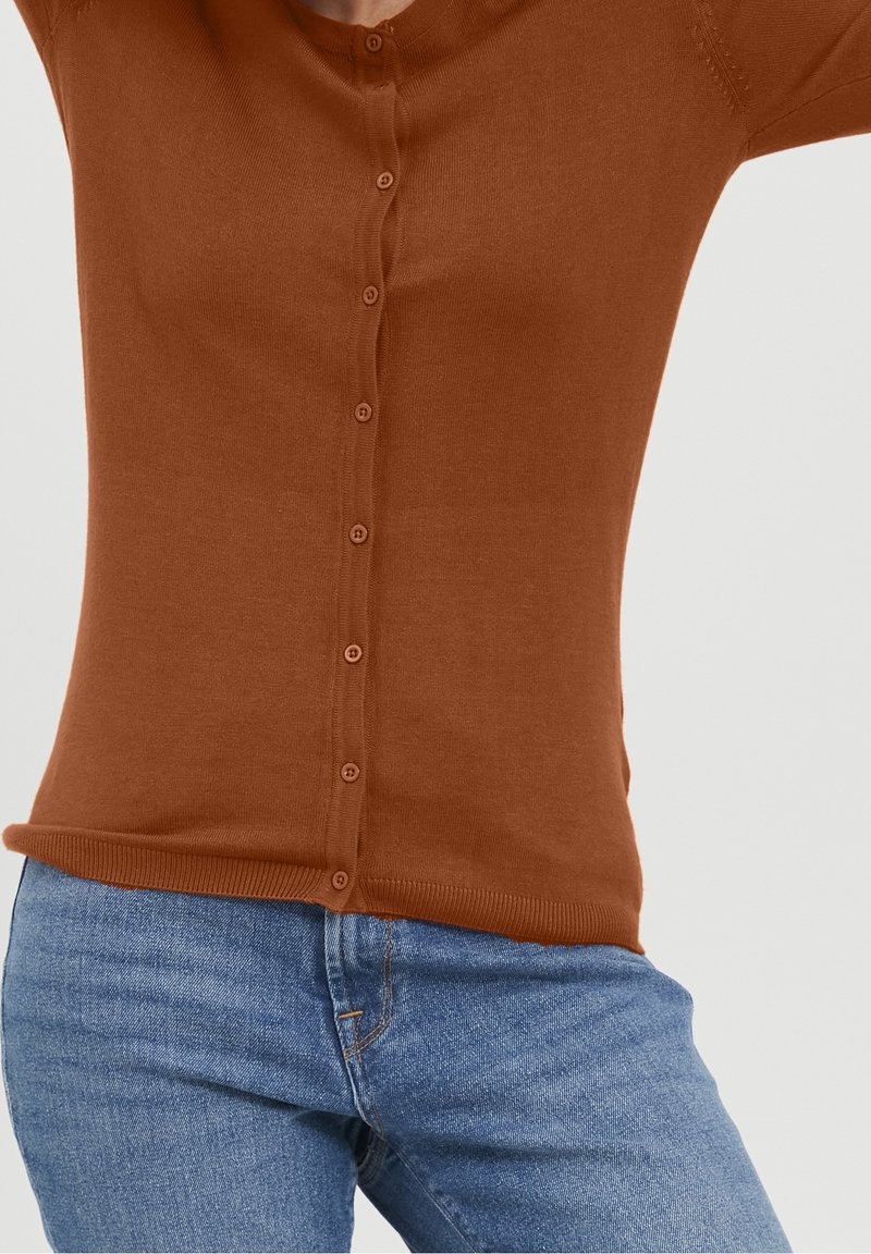 Brun stickad cardigan med knappstängning, ribbat nederkant och långa ärmar, tillsammans med ljusblå jeans. Slät textur, figursydd design.