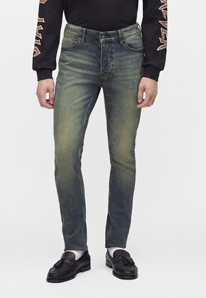 Homme portant un jean bleu slim délavé, des mocassins noirs à pampilles, des chaussettes blanches et une chemise noire à manches longues avec des motifs graphiques sur les manches.