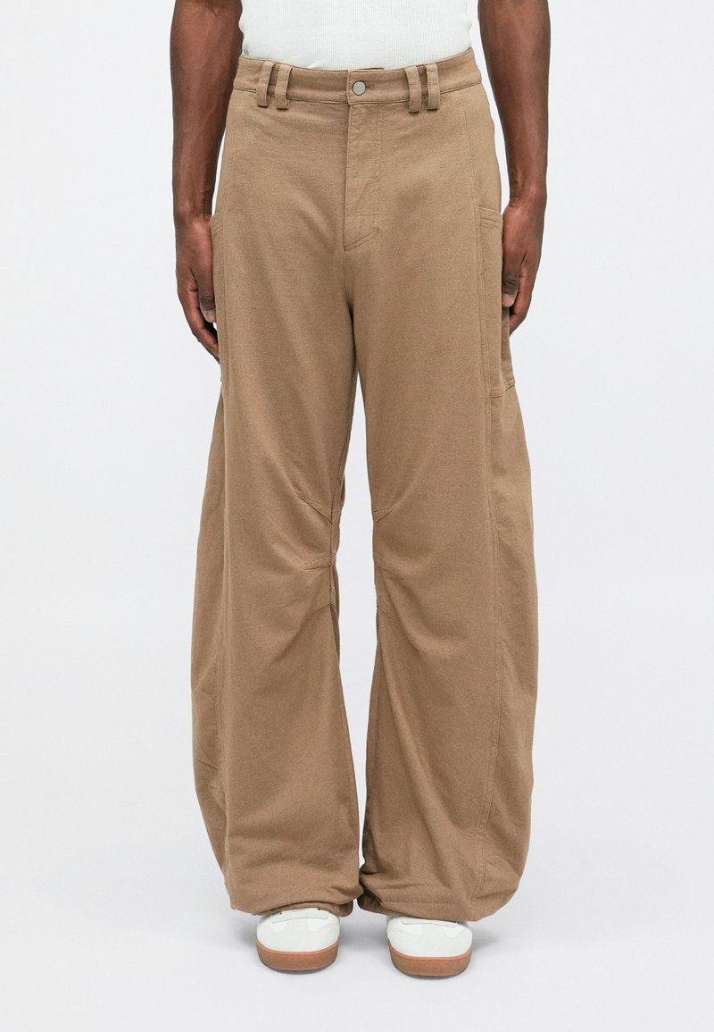 Pantalons beiges amples en tissu texturé, avec des poches latérales, une ceinture avec passants, et une coupe droite se terminant au-dessus des chaussures.