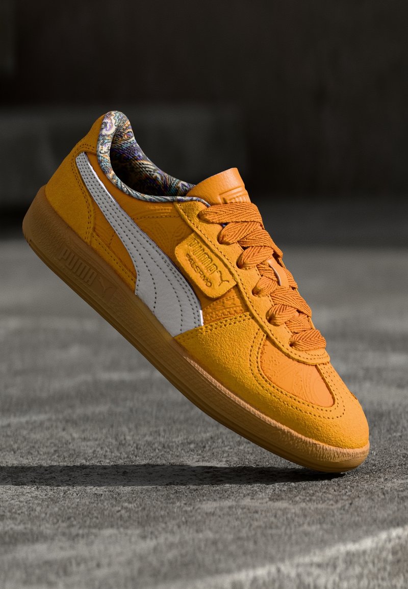 Baskets en daim orange avec une bande Puma blanche, une doublure intérieure à motifs et des lacets, équilibrées sur la semelle contre un fond sombre flou.
