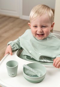 Lichtgroene siliconen voederset bestaat uit een kom met een bijpassende vork en een beker. Heeft kleine witte vlinderontwerpen. Kind zit in een hoge stoel.