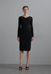 Black long-sleeve bodycon dress with a V-neck, textured fabric, knee-length, paired with black open-toe high-heel shoes.