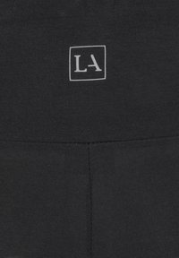 Svarta leggings i elastisk tyg, med en liten vit "LA"-logotyp i en fyrkantig design. Sömmen är synlig längs benen.