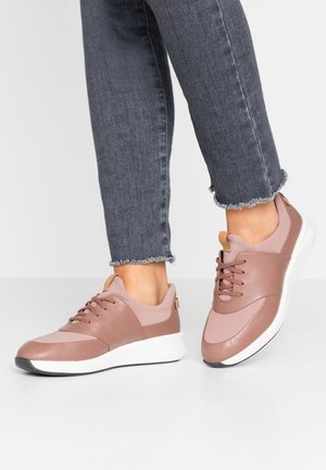 Persoon met roze vetersneakers met witte zolen en grijze jeans met rafelrand op een witte vloer.