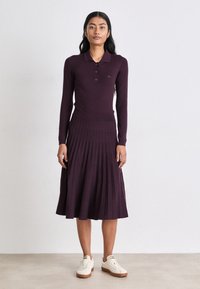 Rebrasta, dugih rukava, tamnoljubičasta haljina s polosuknjom i plisiranom suknjom, uparena s kremastim tenisicama. Glatka tekstura, placket s tri dugmeta.
