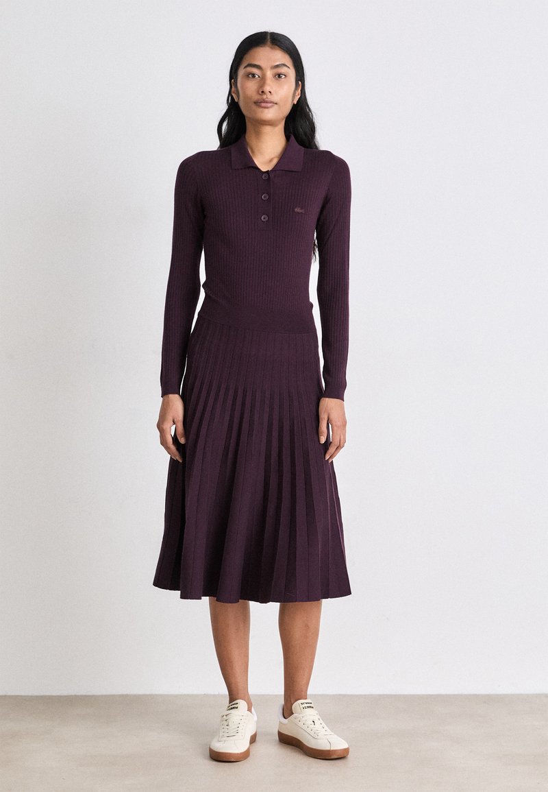 Rebrasta, dugih rukava, tamnoljubičasta haljina s polosuknjom i plisiranom suknjom, uparena s kremastim tenisicama. Glatka tekstura, placket s tri dugmeta.