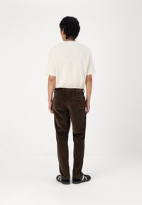 Brune cordfløyelsbukser og en kremfarget kortermet skjorte fra baksiden, iført svarte joggesko med hvite striper.