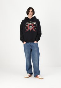 Svart hettegenser med grafisk trykk i rødt og hvitt, kombinert med vide jeans i blå farge og hvite joggesko med røde striper.