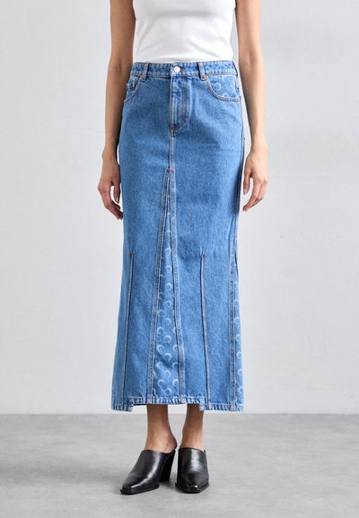 Denim maxiskørt med lige snit, lys blå farve, fremtrædende syninger, forslidser og dekorativt mønster langs nederste kant. Sorte hæle mule-sko.