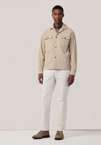 Beige knoopjasje met twee voorzakken, lichte stof, gecombineerd met witte broek en bruine schoenen, met een eenvoudig ontwerp.