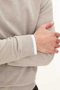 Pull en cachemire beige avec des poignets côtelés, associé à une chemise blanche en dessous, montrant un gros plan du bras et de la main croisés sur le corps.
