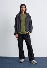Een persoon staat in een casual outfit van een olijfgroen shirt, een grijze jas en zwarte broeken, met sneakers, tegen een effen achtergrond.