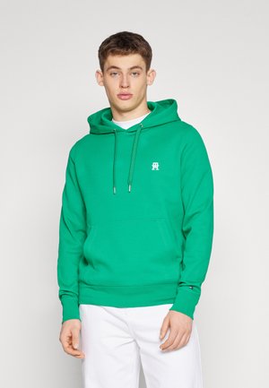 Grüne Hoodie aus weichem Material mit einer Brusttasche, Kapuze mit Kordelzug und kleinem Logo auf der Brust. Kombiniert mit weißen Hosen.