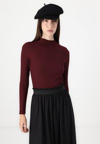 Burgundy ribbad långärmad topp med hög hals i kombination med en svart veckad kjol. Modellen bär en svart basker som accent.