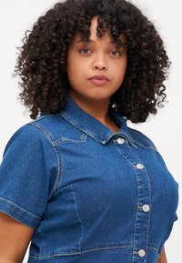 Mørkeblå denimskjorte med korte ermer, krage og sølvfargede trykknapper foran. Har en tettsittende design.