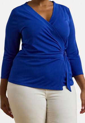 Femme portant un top cache-cœur à manches longues bleu roi et un pantalon beige clair, vue des épaules aux hanches sur un fond uni.