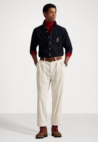 Μπλε πουκάμισο με κουμπιά, κόκκινο ζιβάγκο, κρεμ παντελόνι με πιέτες, καφέ ζώνη και ανθεκτικά καφέ μποτάκια με κόκκινα κορδόνια. Κέντημα στο πουκάμισο.