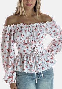 Vrouw draagt een wit off-shoulder blouse met rood bloemenpatroon en veterkorsetdetail, gecombineerd met lichtblauwe jeans.