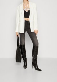 Person som bär en vit kavaj, svart croppad topp med ringdetalj, mörka skinny jeans, svarta knähöga spetsiga stövlar och håller en svart toteväska.