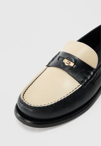 Schwarze und cremefarbene Lederloafer mit glatter Oberfläche, versehen mit einem goldfarbenen Akzent und kontrastierender Naht über dem Zehenbereich.