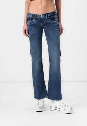 Mellemhøje blå denim bootcut-jeans båret med hvide højt oppe sneakers på en person, der står mod en hvid baggrund.