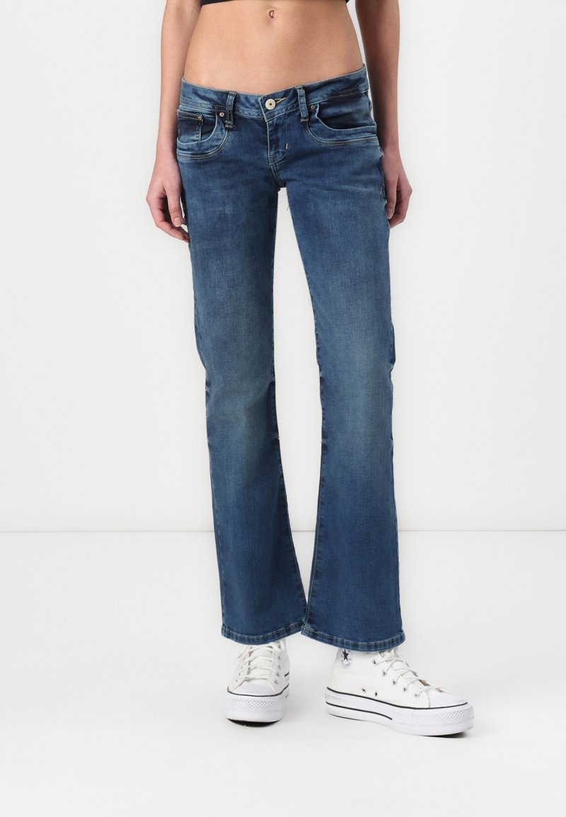 Jean bootcut en denim bleu taille mi-haute, porté avec des baskets blanches montantes sur une personne debout devant un fond blanc.