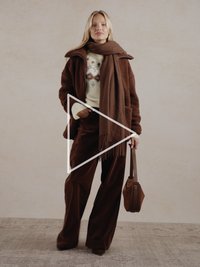 Hnědý strukturovaný oversized kabát, krémový svetr se vzorem, hnědé kalhoty, ladící šála a malá hnědá kabelka. Neutrální pozadí.