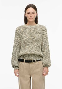 Pull en maille texturée dans des tons beige et vert, avec un col rond et des manches longues légèrement bouffantes. Associé à un pantalon beige taille haute.