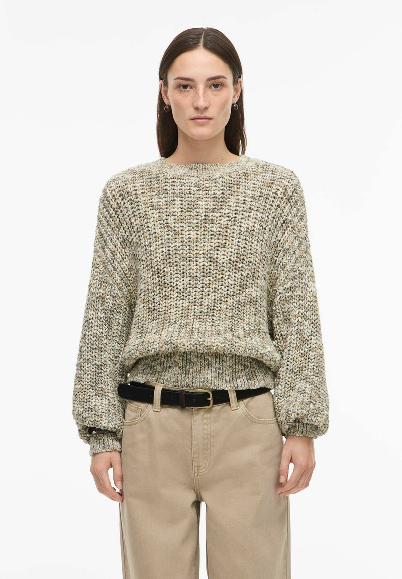 Pull en maille texturée dans des tons beige et vert, avec un col rond et des manches longues légèrement bouffantes. Associé à un pantalon beige taille haute.