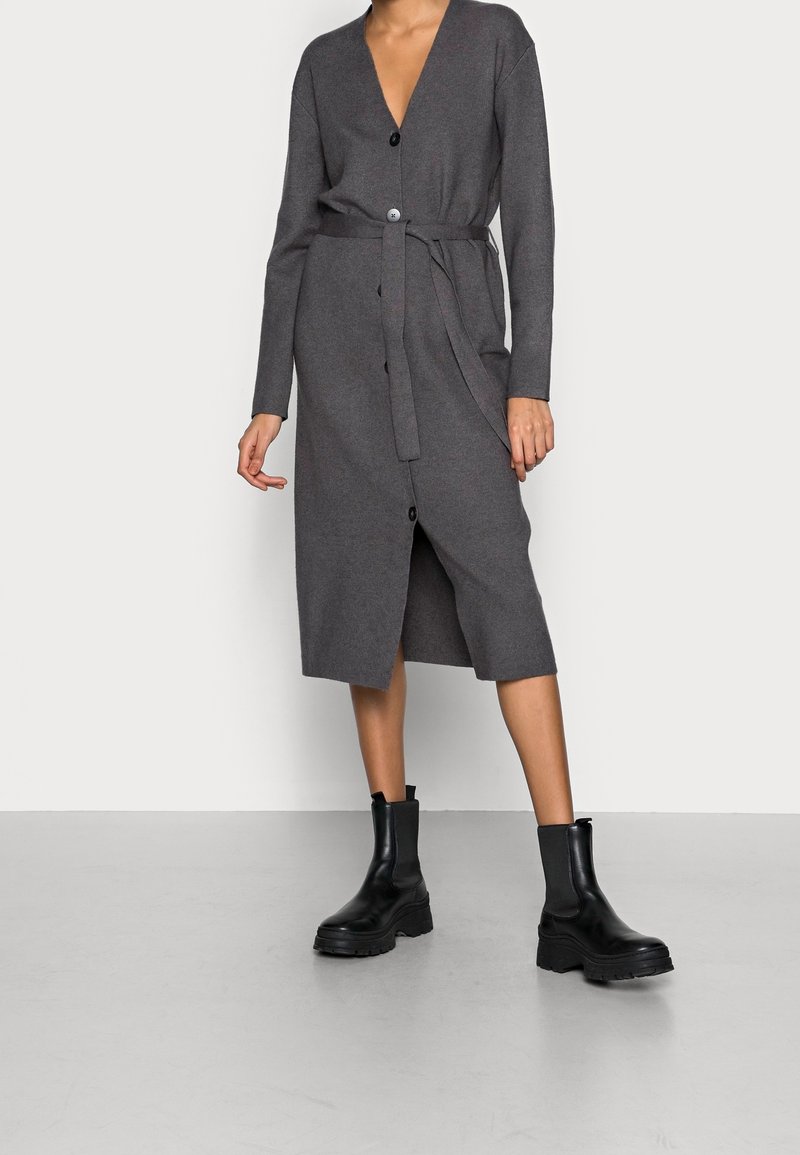 Femme portant une robe cardigan ceinturée gris foncé longueur genou avec des boutons noirs et des bottines noires épaisses, debout sur un sol gris.
