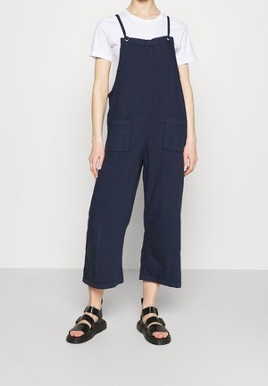 Robe salopette bleu marine en tissu respirant, avec jambes larges, bretelles réglables et deux poches avant. Associée à des sandales noires.