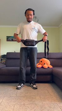 Μαύρη μέση τσάντα κατασκευασμένη από συνθετικό υλικό, με λεία επιφάνεια και μπροστινό καπάκι. Ορατά καστανά τζιν και μαύρα αθλητικά παπούτσια.