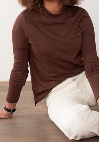 Brown long-sleeve top made from a soft, stretchy material with a mock neck; paired with white pants; smooth texture and relaxed fit.