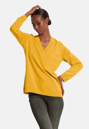 Gelber V-Ausschnitt-Strickpullover mit langen Ärmeln und geripptem Saum, kombiniert mit dunkelgrünen Hosen. Weiche Textur, lockere Passform.