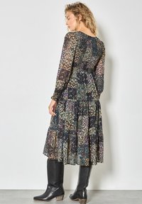 Abito maxi con maniche lunghe, tessuto multi-patternato in blu scuro, verde e oro, con vita arricciata e gonna a strati. Stivali neri fino al polpaccio.