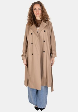 Donna con capelli ricci che indossa un trench beige doppiopetto, jeans blu a gamba larga e scarpe nere con la punta, in piedi davanti a uno sfondo neutro.