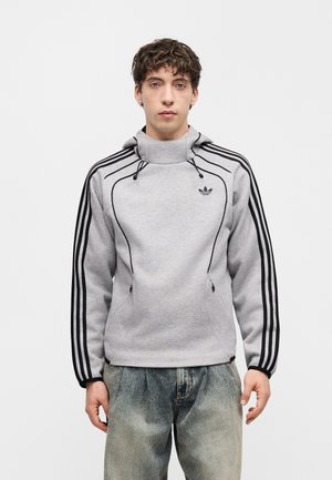 Młody mężczyzna w szarej bluzie z kapturem Adidas z czarnymi paskami wzdłuż rękawów i wysokim kołnierzem, w połączeniu z wyblakłymi niebieskimi dżinsami.