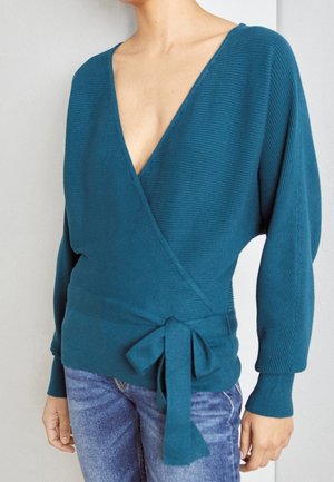 Woman wearing teal wrap sweater with deep V-neck and tied waist, paired with blue jeans, standing against a plain background.