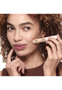 Frau mit lockigem Haar, die den KIKO Milano Love Fusion Concealer nahe ihrer Wange hält, trägt ein braunes ärmelloses Oberteil und lächelt leicht.