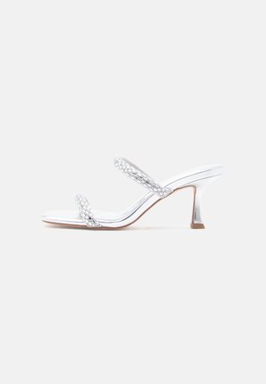Silver heeled sandals feature two beaded straps, a pointed toe, and a mid-height, block heel. Smooth, shiny material with a textured accent.