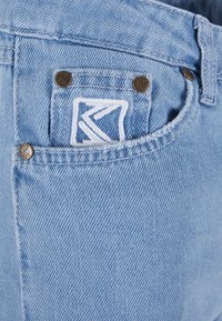 Svetlomodré džínsy majú hnedý kovový hardvér, logo nášivku s bielym vyšívaním na zadnej vrecku a textúrovaný povrch látky.