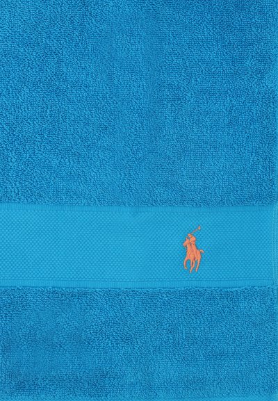 Φωτεινή μπλε πετσέτα με υφασμάτινη λωρίδα κοντά σε μία άκρη και μικρό πορτοκαλί κεντημένο λογότυπο παίκτη πόλο πάνω σε άλογο.