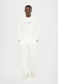 Fehér pamut pulóver és hozzá illő nadrág "Armani Exchange 1991" logóval. A modellen bézs sportcipő van. Egyszerű, letisztult dizájn és puha textúra.