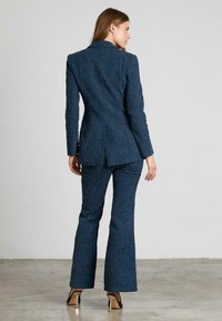 Traje texturizado azul marino que presenta una blazer entallada con una abertura y pantalones acampanados, combinados con zapatos de tacón alto sobre un fondo neutro.