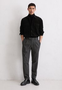 Uomo in piedi contro un muro semplice, vestito con una camicia nera con bottoni, pantaloni grigi e scarpe nere stringate, con le mani nelle tasche.