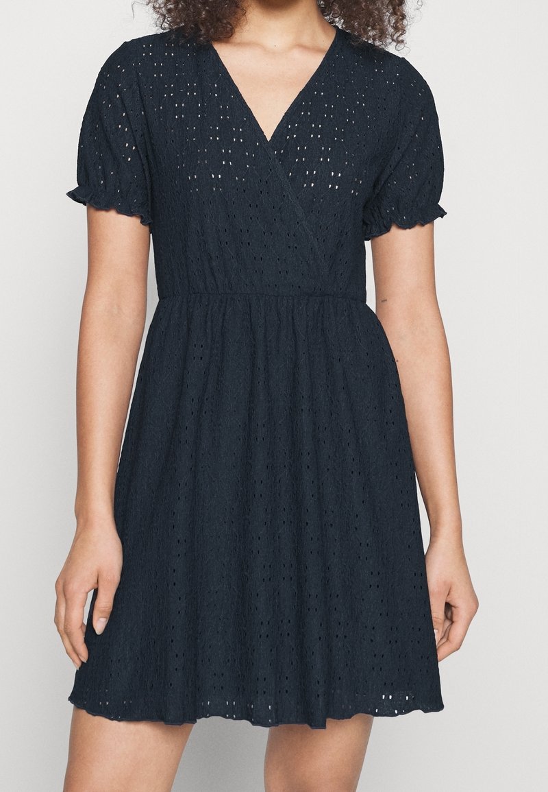 Femme portant une robe bleu marine à manches courtes en œillet avec un décolleté en V et une taille froncée, debout devant un fond clair uni.