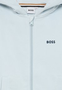 Hoodie azul claro com fecho, feito de tecido suave, apresentando um pequeno logotipo "BOSS" em azul marinho, bolsos frontais e capuz forrado.