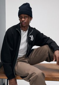 Giacca nera con zip e logo bianco, t-shirt grigia, pantaloni marroni e berretto nero a costine. Modello seduto su una panchina di legno.